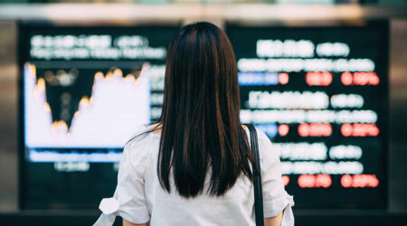 Woman in white shirt with long brown hair standing and facing electronic boards with graphs and data displayed, but blurred.