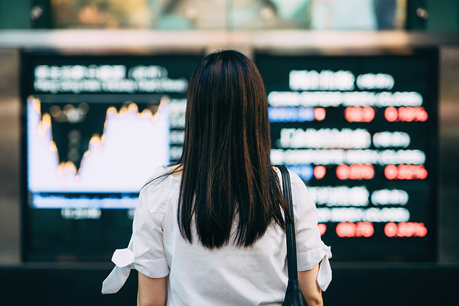 Woman in white shirt with long brown hair standing and facing electronic boards with graphs and data displayed, but blurred.